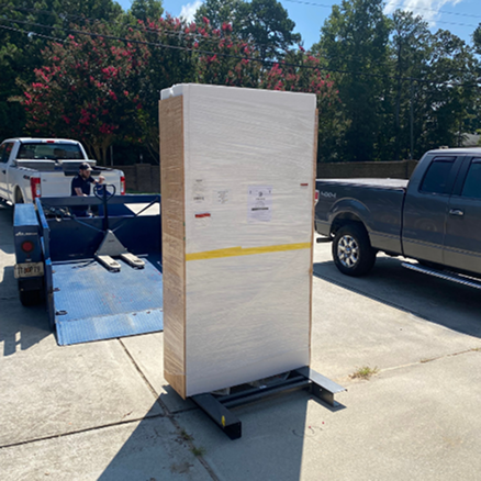 Large wooden pallet with a white label on a concrete surface with vehicles and trees in the background.