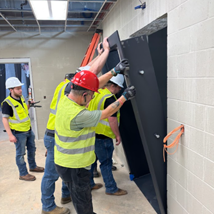 Workers installing a large black panel on a wall in an indoor setting.