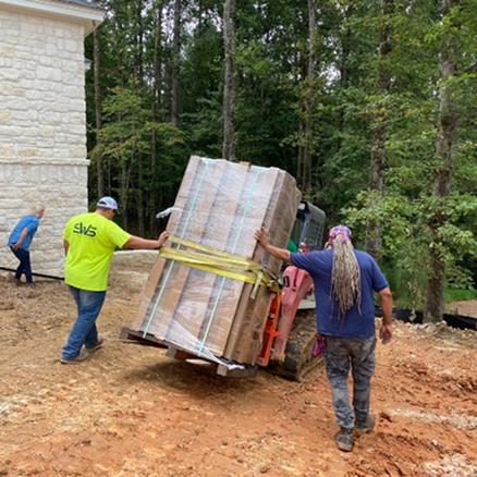 Two men moving a large pallet of bricks with a tractor in a wooded area.