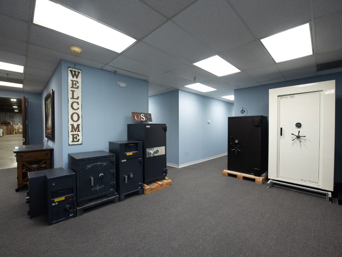 A showroom or storage room displaying multiple heavy-duty safes. On the left, a vertical white 'WELCOME' sign hangs on a light blue wall above a row of black safes on wooden pallets.