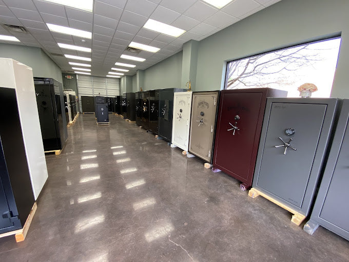 A row of various gun safes in different colors including gray, burgundy, beige, and black, displayed in a showroom with a concrete floor and large windows.