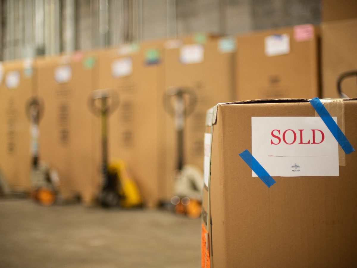 A brown cardboard shipping box in the foreground with a white 'SOLD' label taped on with blue tape, set in a warehouse with other boxes and pallet jacks blurred in the background.