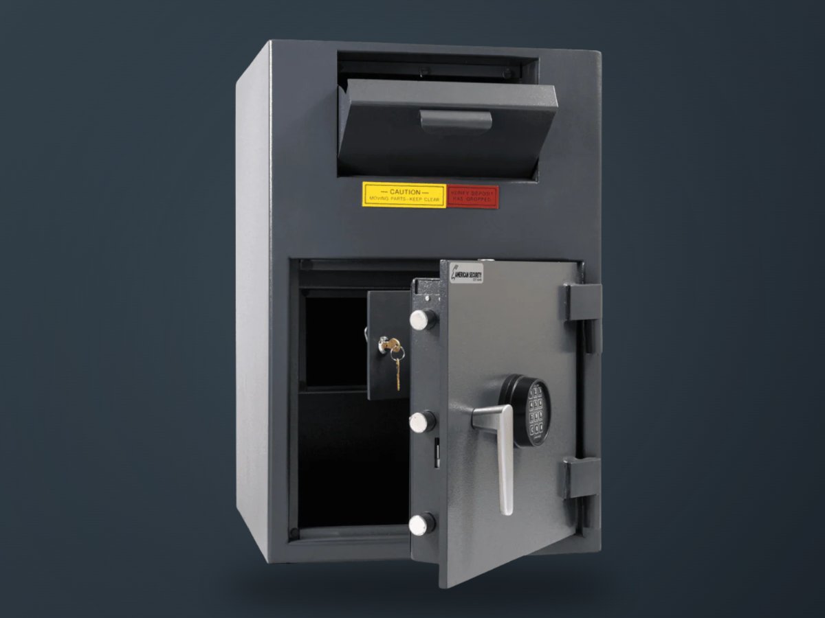 A gray metal safe with an open door, showing a lock, keypad, and interior compartment. The safe has a top cash drawer with a pull tab and a yellow warning label on a dark gray background.