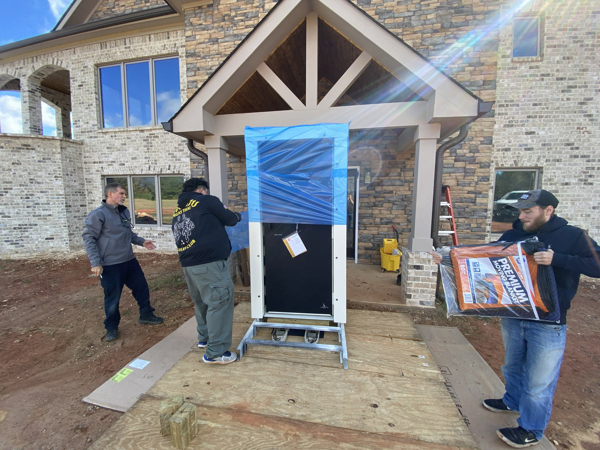 Delivery crew unloads a large safe at a home entrance, using protective wrap and floor covering—showing what happens on safe delivery day.