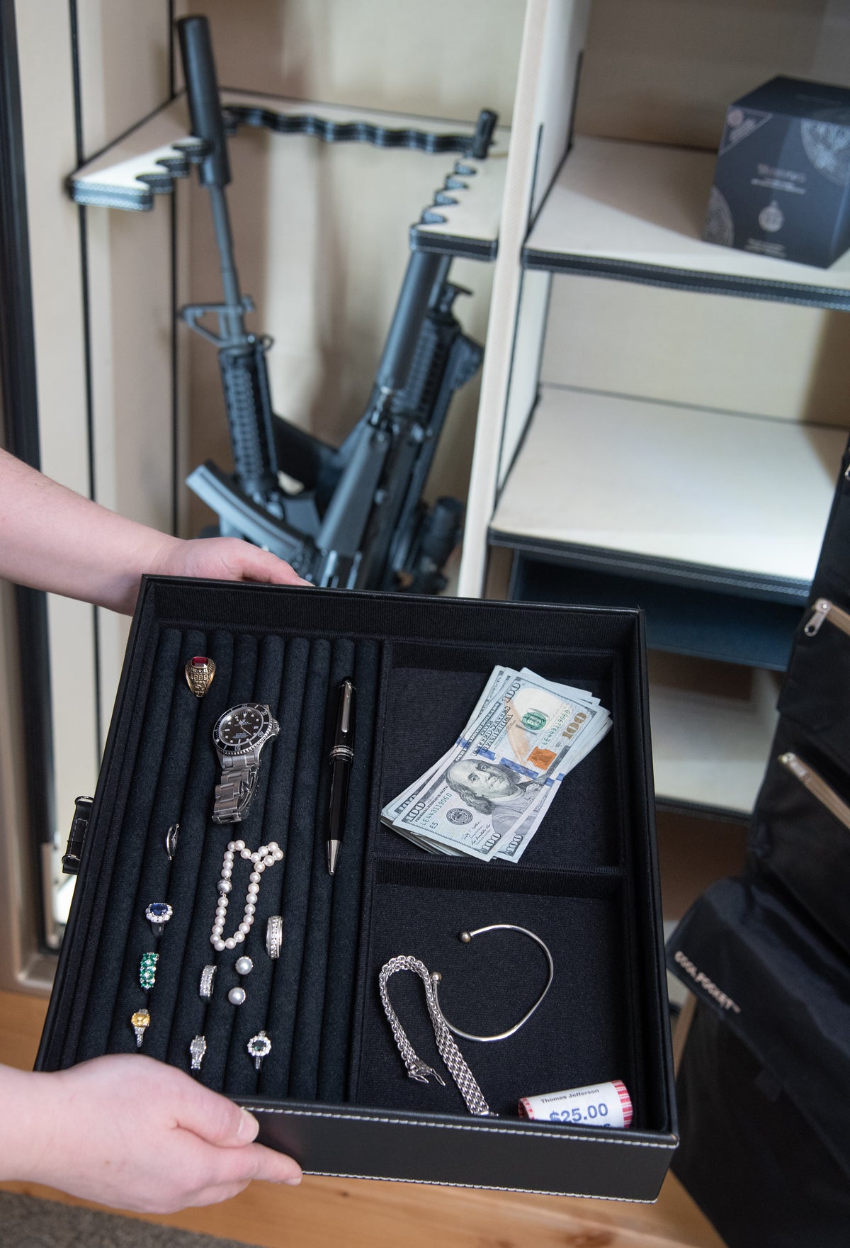 Hands hold a tray of jewelry, a watch, and cash in front of an open safe with firearms and shelving in the background.