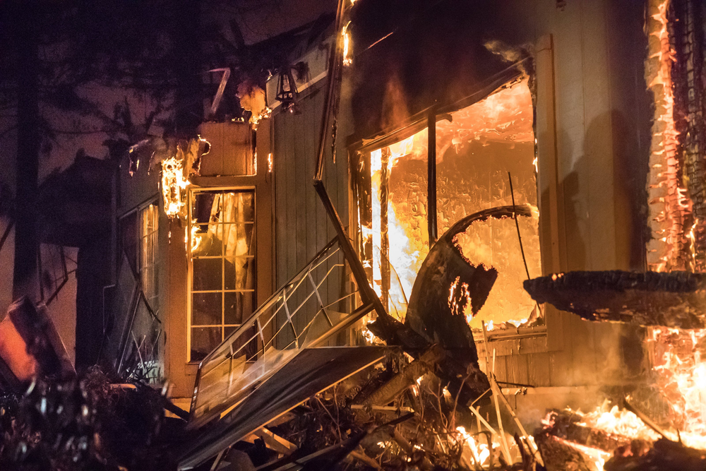 House engulfed in flames at night, with fire visible through windows and along the exterior walls.
