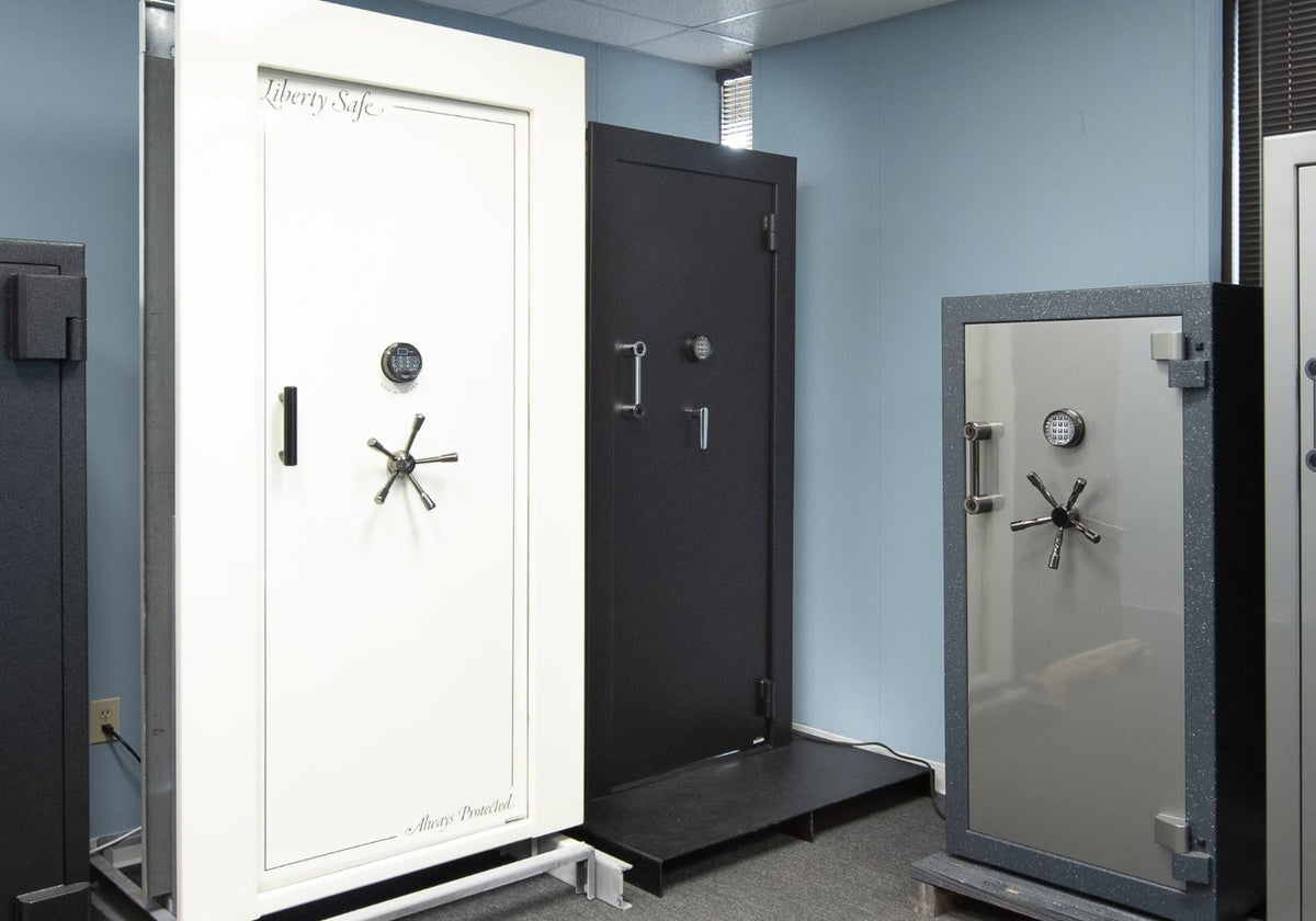 Three heavy-duty security safe doors displayed side-by-side in an indoor showroom. The left safe is white with a black handle and dial; the middle safe is matte black; the right safe has a gray textured frame and a clear front door.