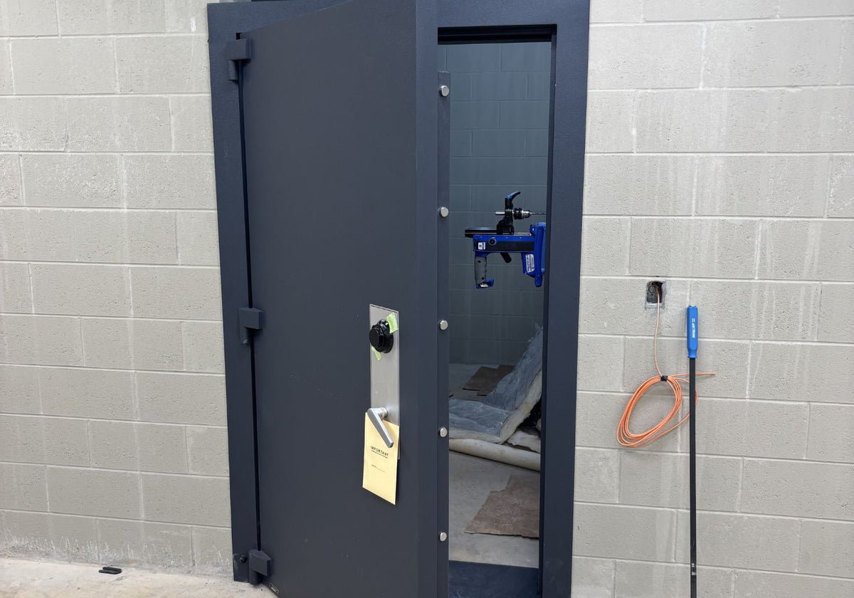 A dark gray industrial security door with a handle and a yellow tag, set against a light gray cinderblock wall. A blue tool is mounted on the door, and an orange coiled hose is visible on the right side.