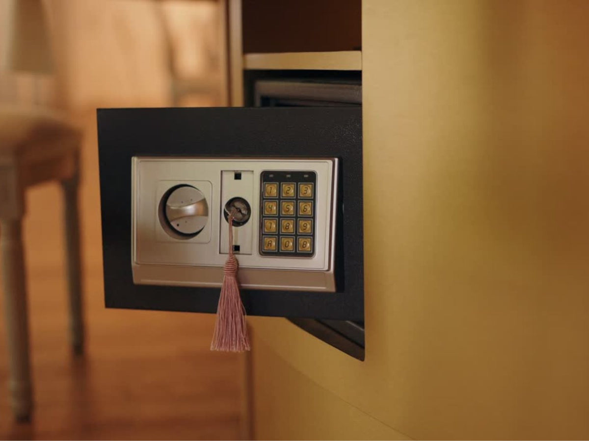 A black electronic safe with a silver keypad, dial lock, and a pink tassel key holder, shown mounted on a wall in a warm indoor setting.