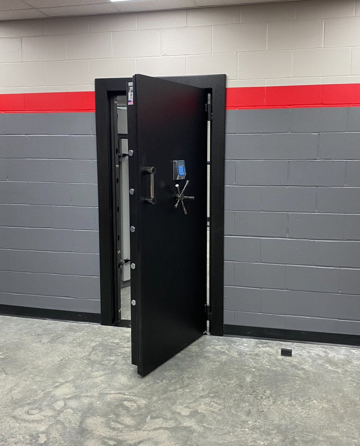 A heavy-duty black metal safe or security door with a digital keypad lock and crossbar handle, shown slightly open in a tiled room with gray block walls and a red stripe.